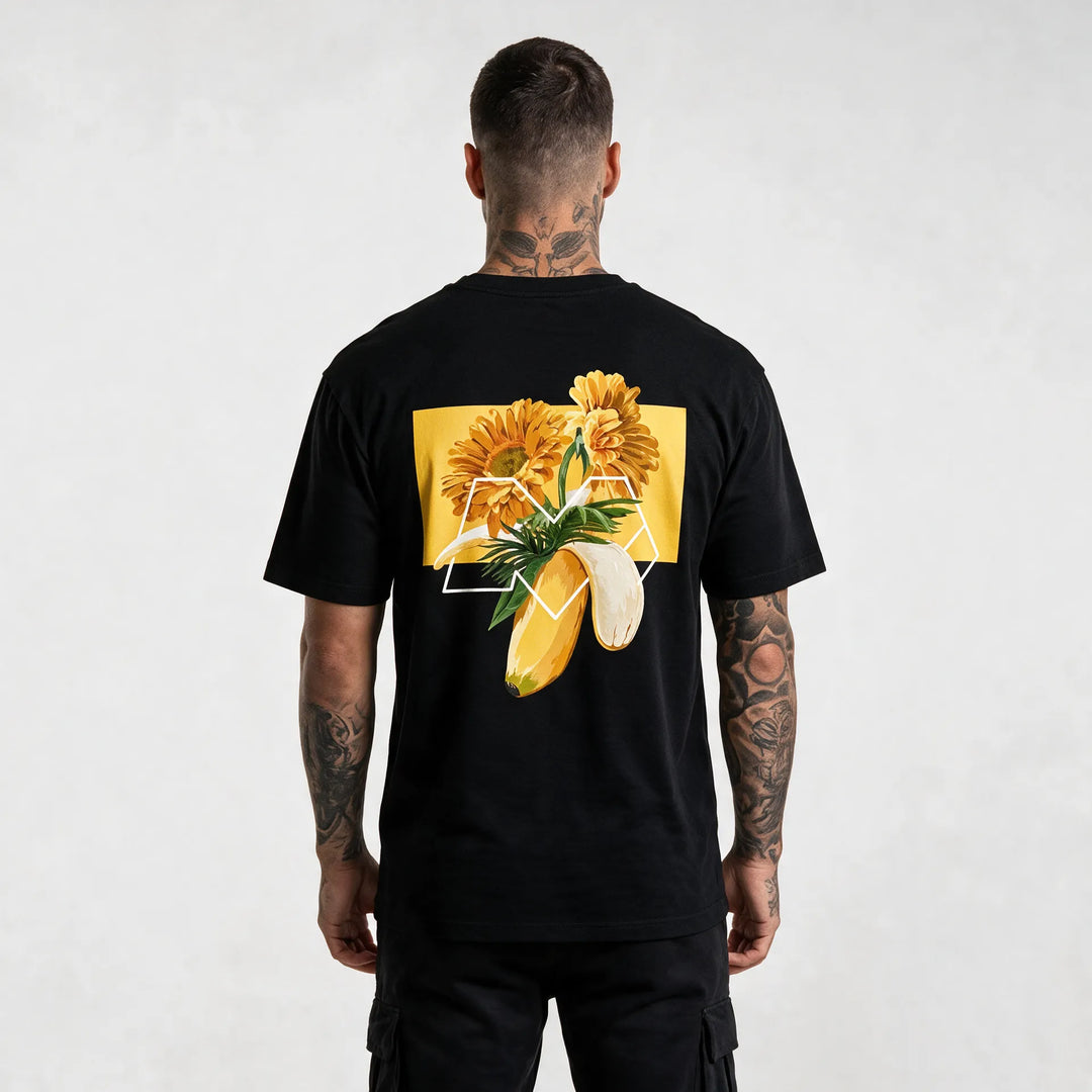 Back of a black banana t-shirt with orange gerberas, palm leaves and a white M outline on a yellow block