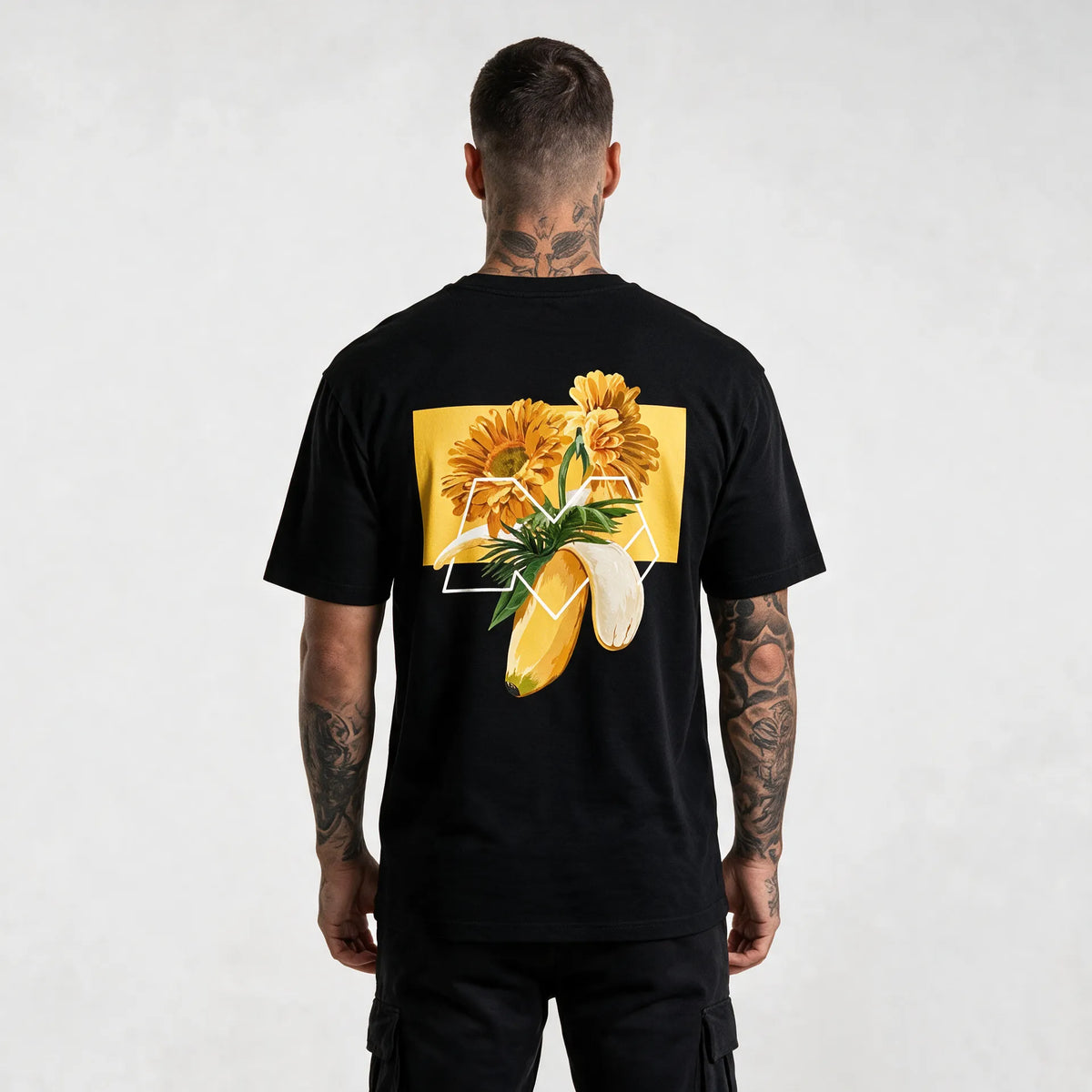 Back of a black banana t-shirt with orange gerberas, palm leaves and a white M outline on a yellow block