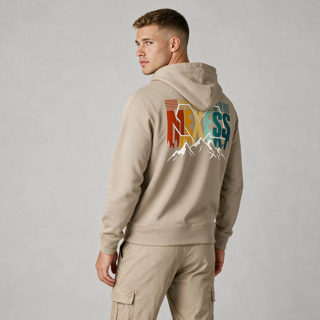 Man wearing a beige mountain hoodie, back view of the rust, yellow and teal MEXESS lettering with white peaks